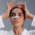 A woman wearing a white pajama shirt holds her hands to her temples while wearing a white adhesive nose patch against a solid gray background.